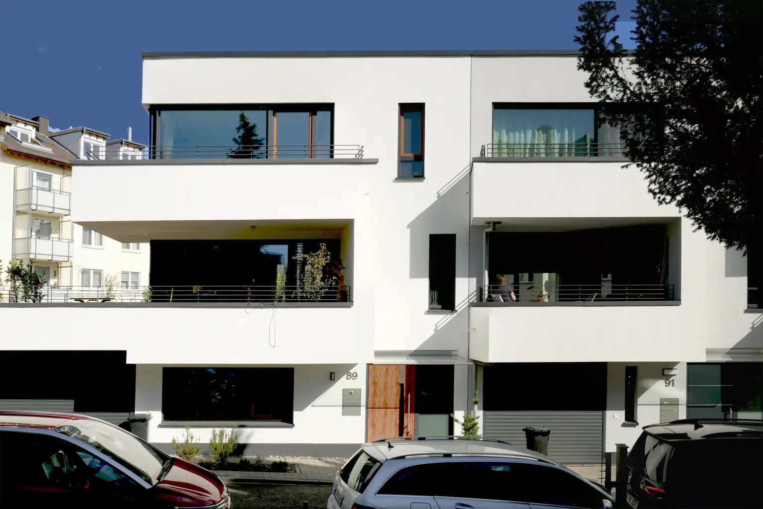 Frontansicht eines modernen, weißen Mehrfamilienhauses mit großzügigen Balkonen, dunklen Fensterrahmen und Garagen im Erdgeschoss.