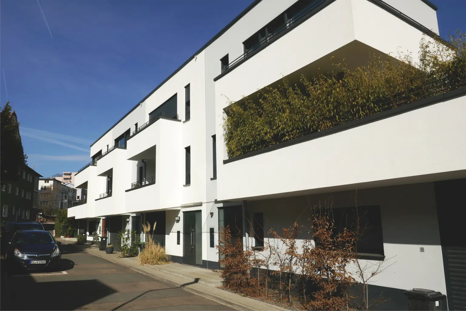 Straßenseite einer modernen Wohnanlage mit mehreren Wohneinheiten, Balkonbepflanzung und geparkten Autos in einer ruhigen Seitenstraße.