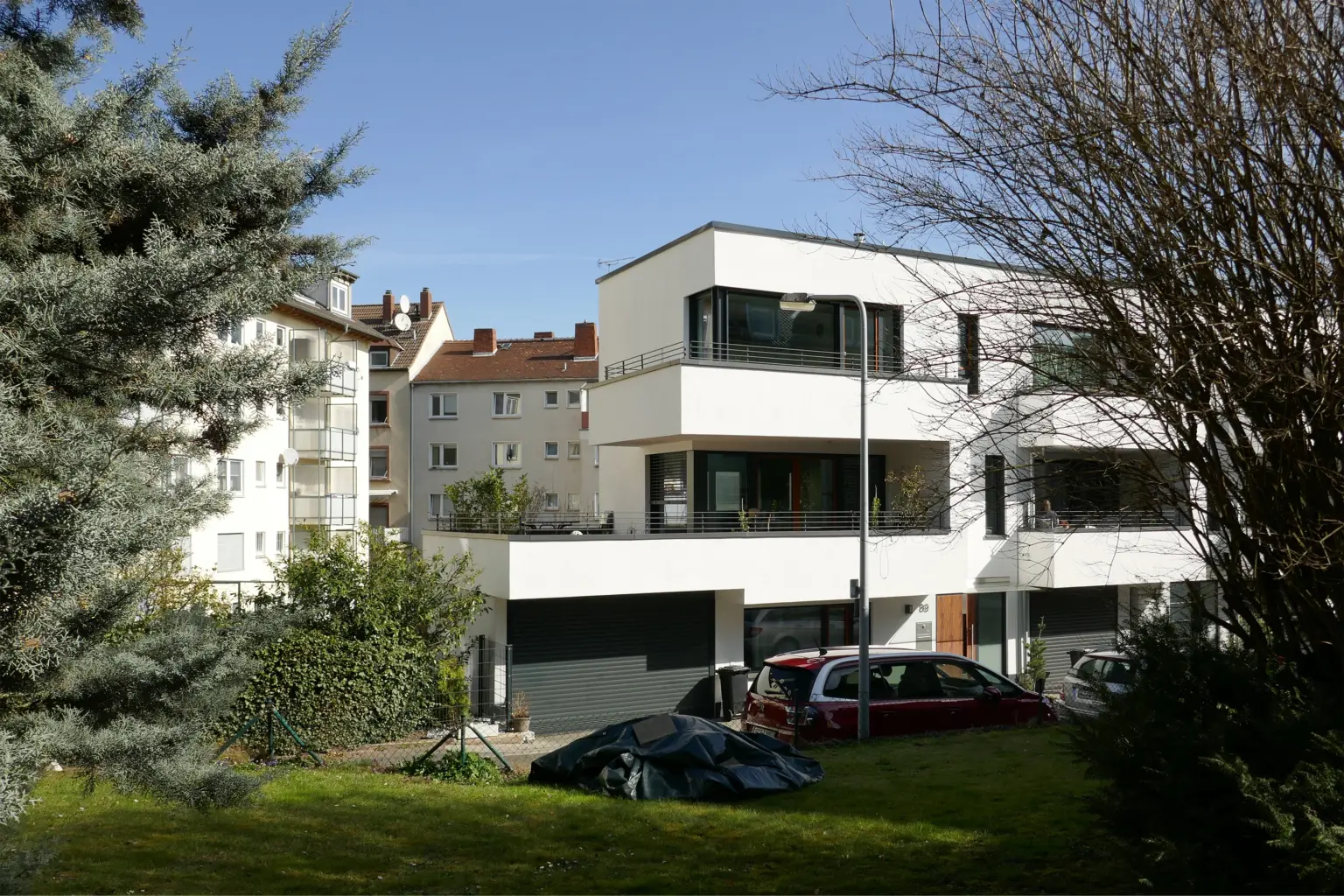 Blick durch Bäume auf ein modernes Wohngebäude in einem urbanen Viertel mit Kontrast zwischen Neubau und Bestandsarchitektur im Hintergrund.