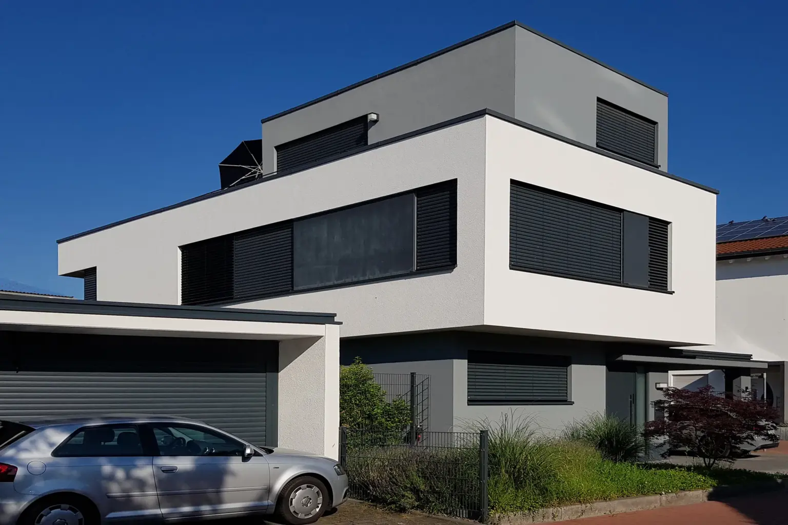 Außenansicht eines modernen Gebäudes mit weißer Fassade, anthrazitfarbenen Akzenten und geschlossenen Außenjalousien bei hellem Tageslicht.