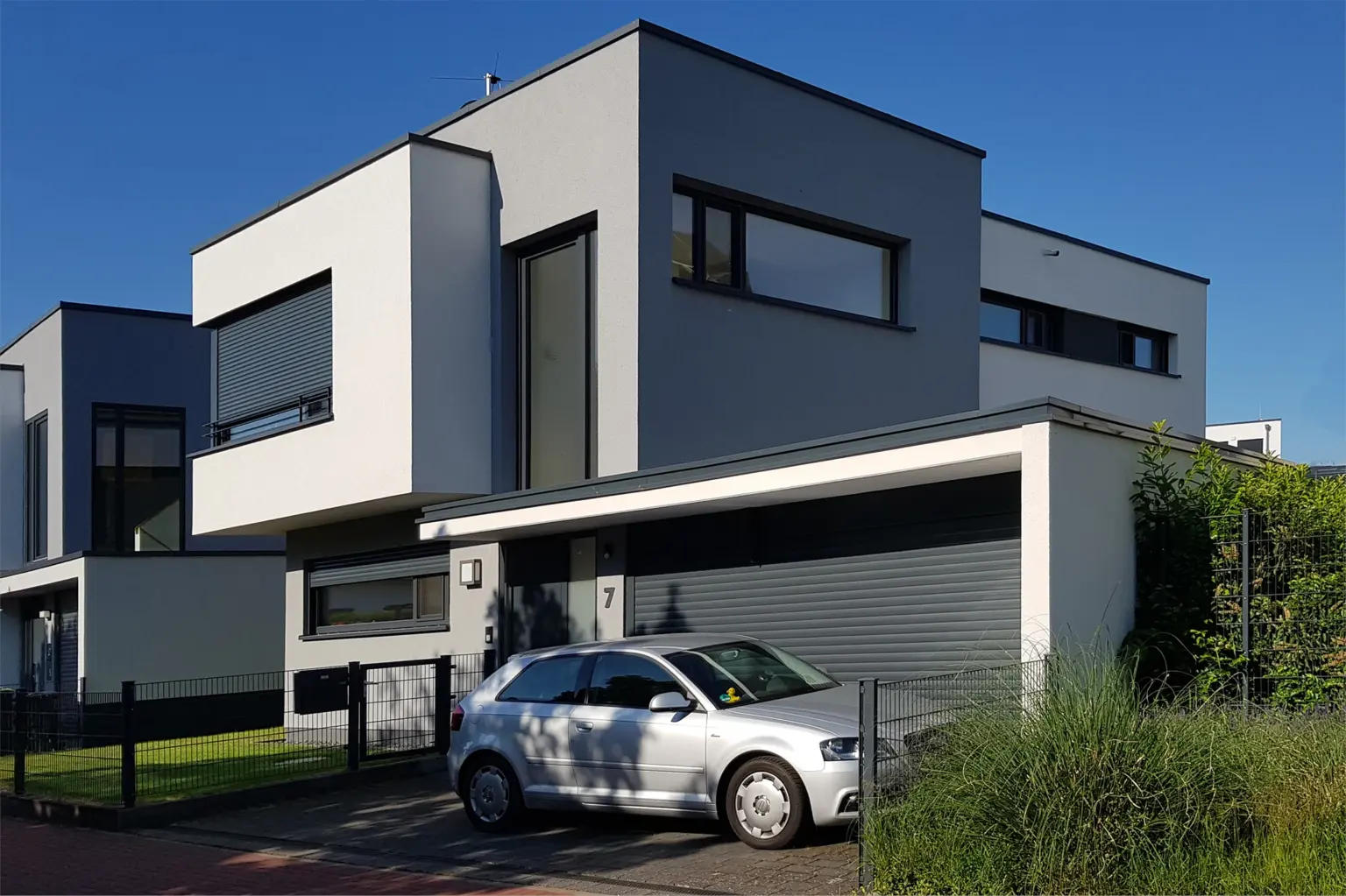 Moderne Architektur: Zweigeschossiges Wohnhaus in Weiß und Grau mit markanten Fensterfronten und einem silbernen Auto in der Einfahrt unter blauem Himmel.