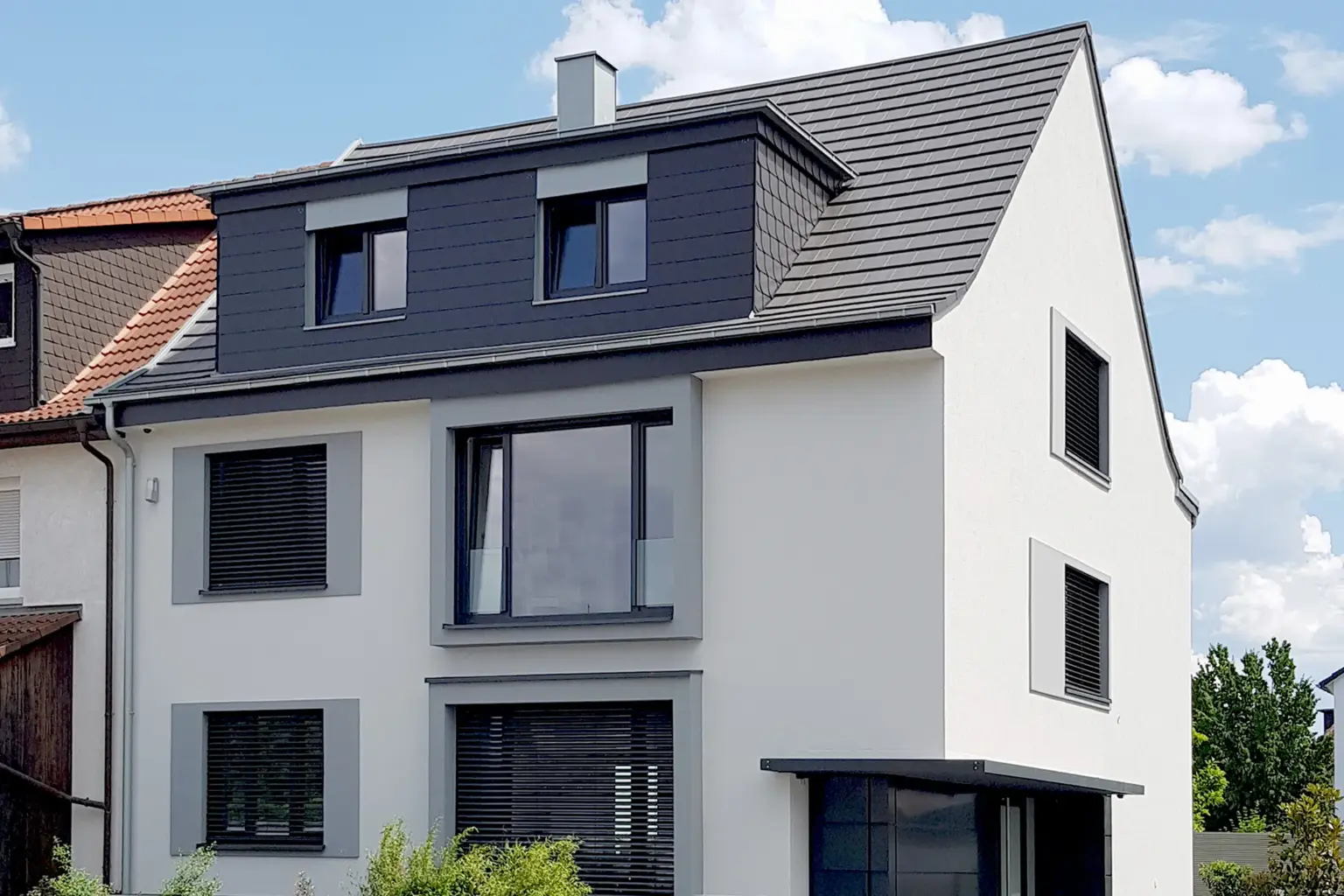 Straßenansicht eines modernen Hauses mit weißer Fassade, grauen Fensterumrandungen und einem anthrazitfarbenen Satteldach mit Gauben.