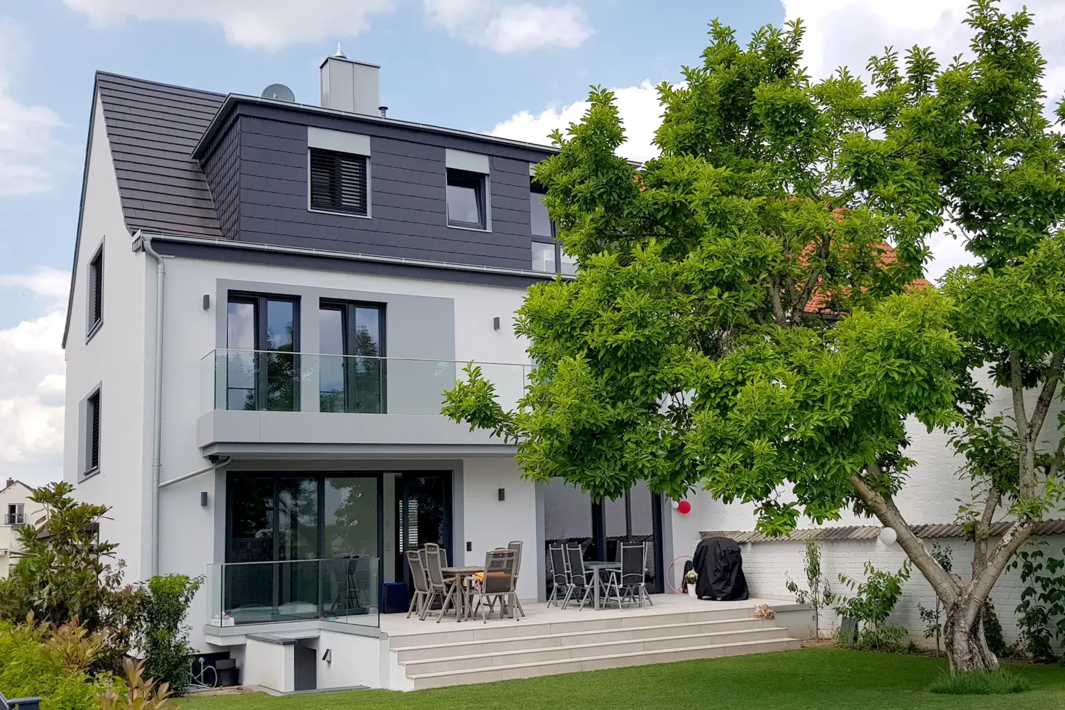 Gartenansicht eines modernen Einfamilienhauses mit großer Terrasse, gläsernen Balkonbrüstungen und einem großen, schattenspendenden Baum.