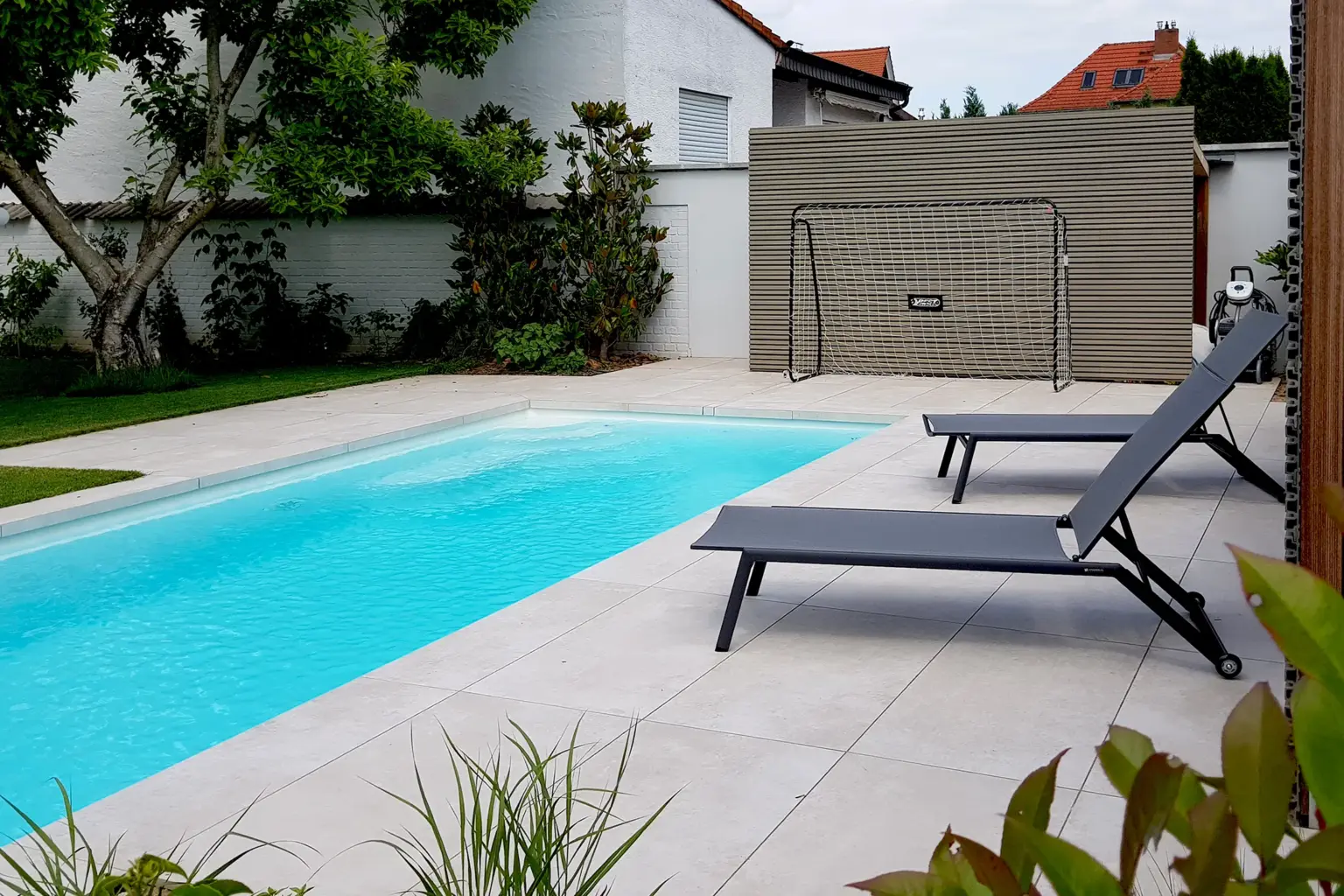 Moderner Außenpool mit hellen Fliesen, zwei dunklen Sonnenliegen und einem Fußballtor im Hintergrund auf einer gepflegten Rasenfläche.