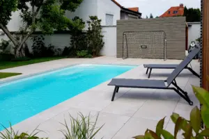 Moderner Außenpool mit hellen Fliesen, zwei dunklen Sonnenliegen und einem Fußballtor im Hintergrund auf einer gepflegten Rasenfläche.