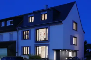 Nachtaufnahme eines beleuchteten modernen Hauses; die warmen Lichter hinter den Jalousien erzeugen eine wohnliche Atmosphäre.