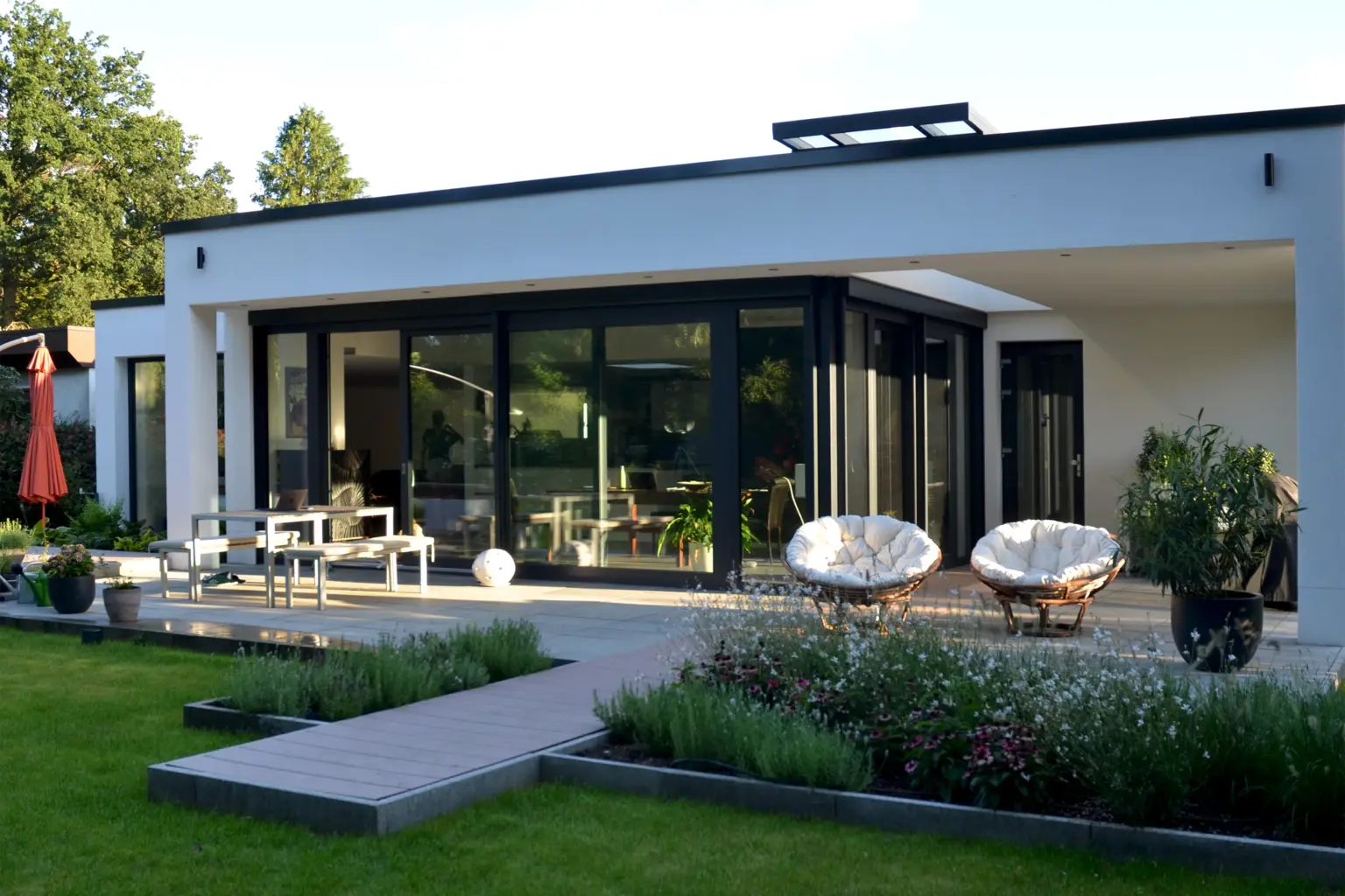 Große Glasfront eines modernen Bungalows bei Abenddämmerung mit Blick auf die Terrasse, Sitzgruppe und gemütliche Papasan-Sessel.