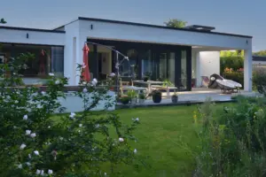 Gartenperspektive mit blühenden Hibiskussträuchern im Vordergrund und Blick auf die offene Architektur des Hauses mit Sonnenschirm.