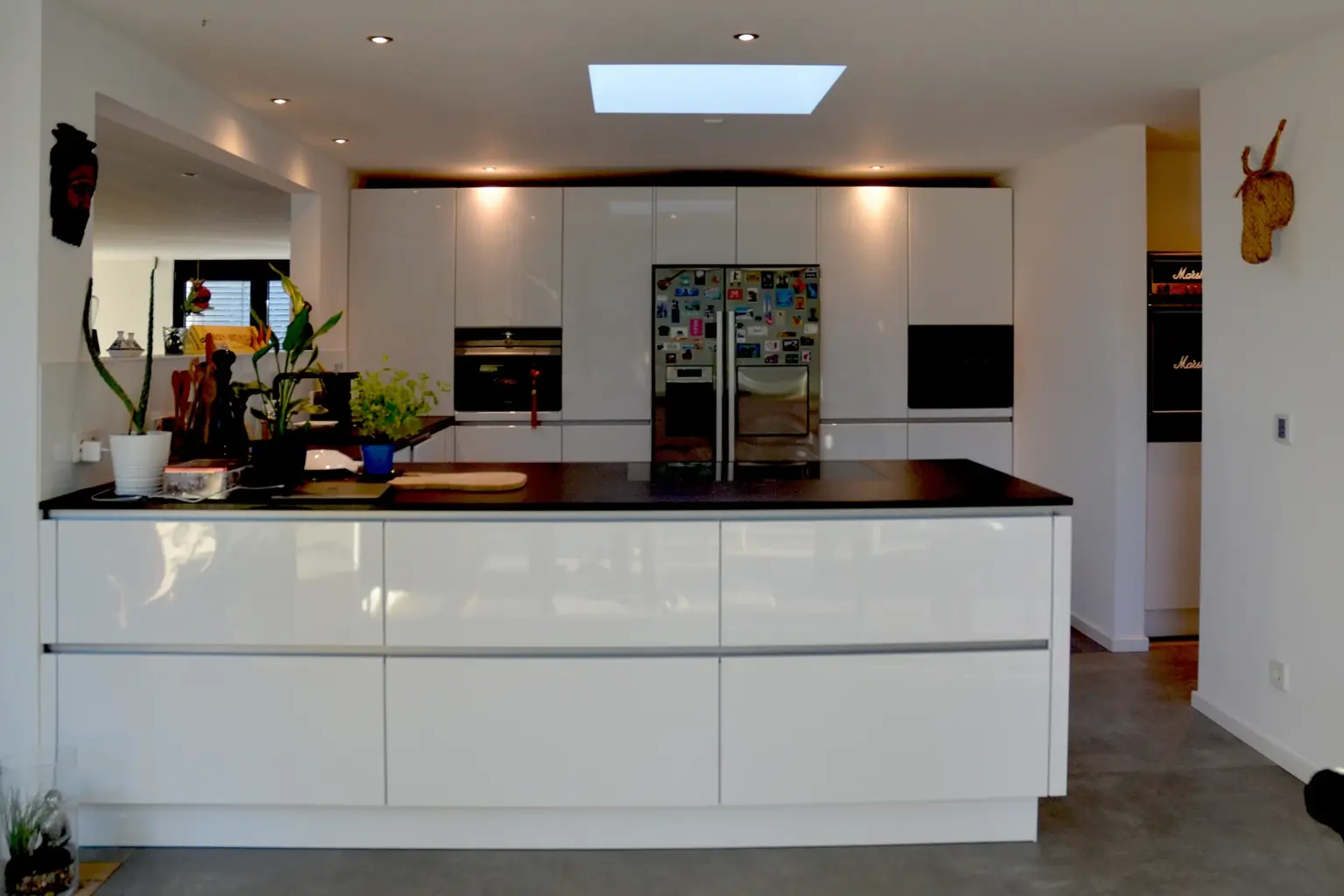 Moderne, minimalistische Küche in Hochglanz-Weiß mit Kochinsel, schwarzer Arbeitsplatte und einem Oberlicht für natürliches Tageslicht.