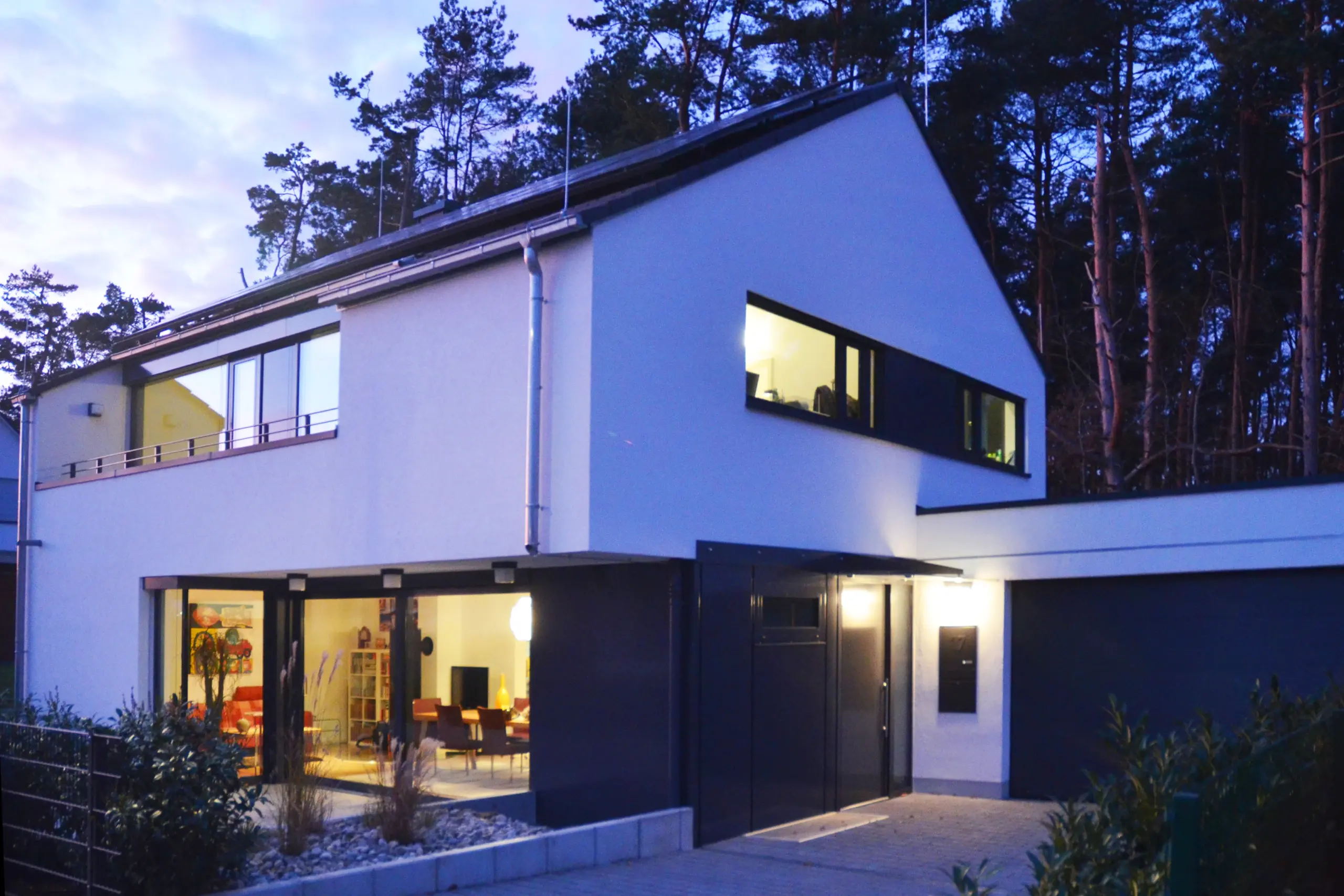 Dämmerungsaufnahme eines modernen Hauses am Waldrand mit beleuchteten Fenstern, asymmetrischem Pultdach und einer integrierten Garage.