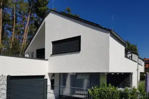 Hell erleuchtete Außenansicht eines weißen Wohnhauses mit markanter Dachschräge, schwarzen Raffstores und einem modernen Metallzaun vor blauem Himmel.