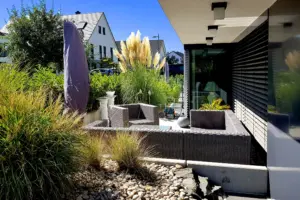 Gemütliche Lounge-Ecke auf einer Terrasse mit grauen Rattanmöbeln, umgeben von Gräsern, Pampasgras und einer modernen Kiesgestaltung.