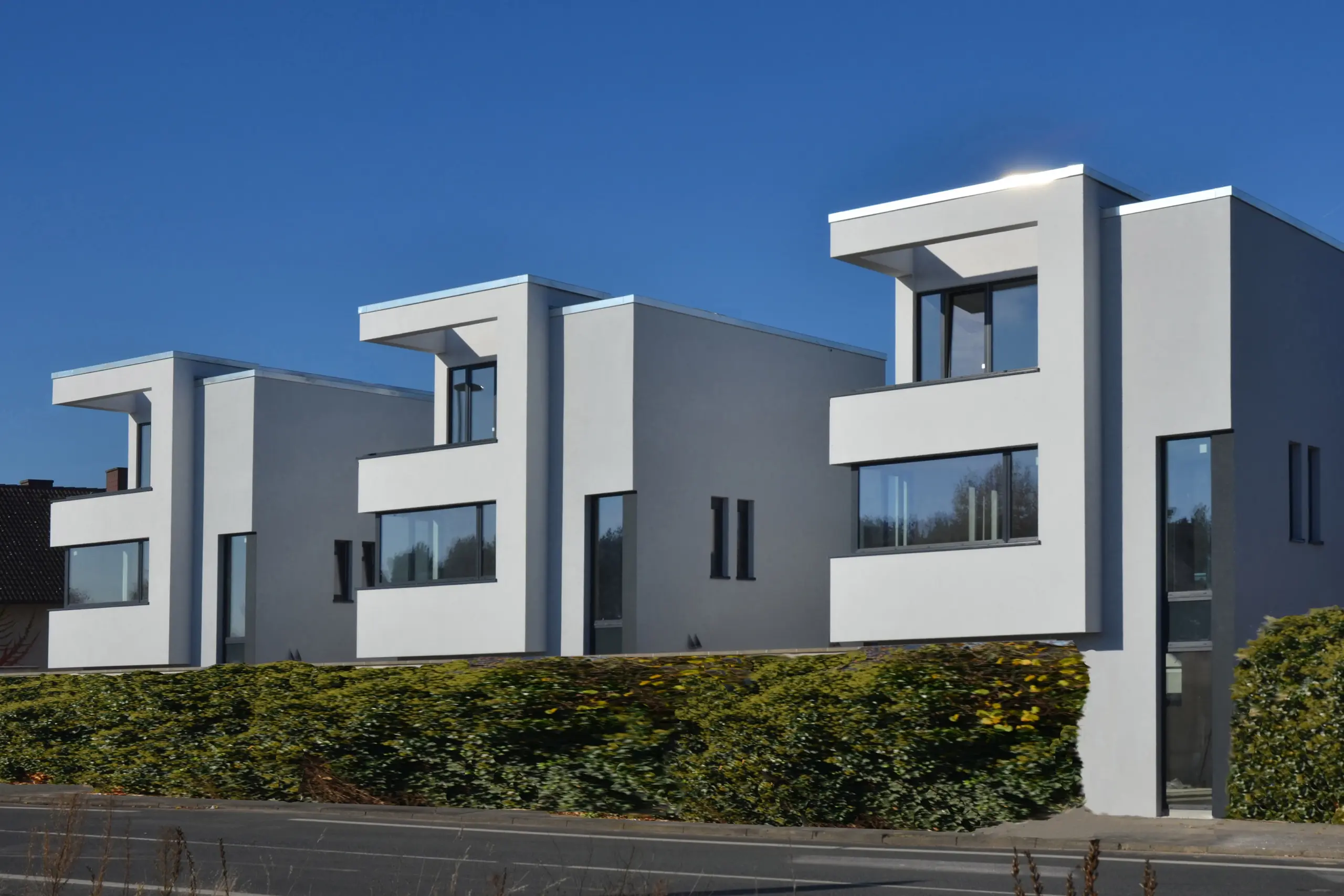 Drei identische, moderne Stadthäuser in kubischer Bauweise mit weißer Fassade und Übereck-Fenstern hinter einer hohen grünen Hecke.