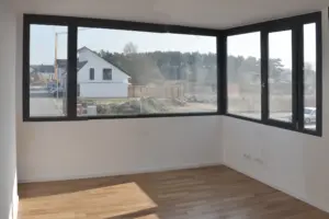 Innenansicht eines lichtdurchfluteten, leeren Raums im Neubau mit großem Eckfenster, Parkettboden und Blick ins Grüne.