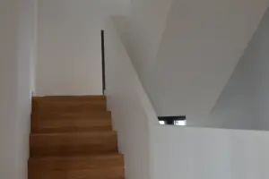 Minimalistisches Treppenhaus in einem modernen Wohnhaus mit einer Holztreppe und schlichten weißen Wandflächen.