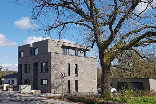 Dreigeschossiges modernes Wohnhaus mit grauer Klinkerfassade und einem zurückgesetzten Staffelgeschoss unter blauem Himmel mit markantem altem Baum im Vordergrund.