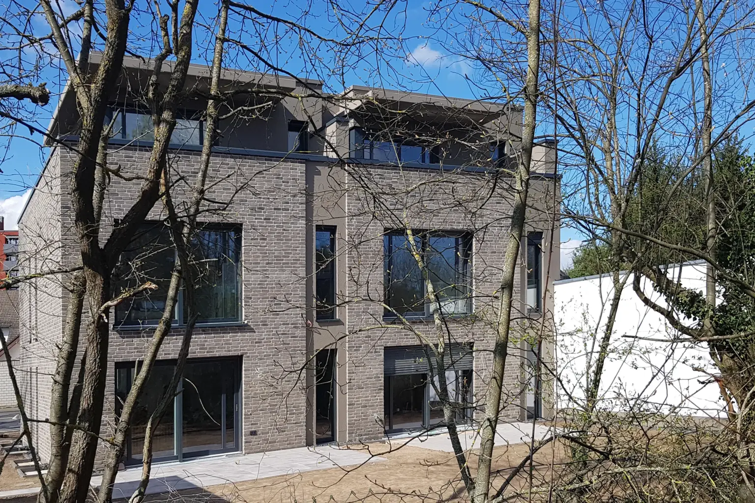 Rückansicht des Klinkergebäudes mit großen Glasschiebetüren zur Terrasse, fotografiert durch die Äste eines Baumes bei hellem Tageslicht.