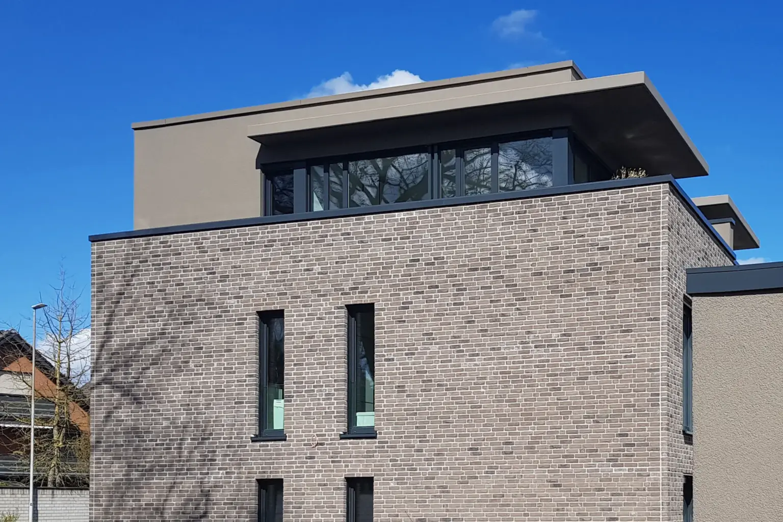 Detailaufnahme des obersten Stockwerks mit Fokus auf das flache Dach und die schmalen, vertikalen Fensteröffnungen in der Backsteinfassade.