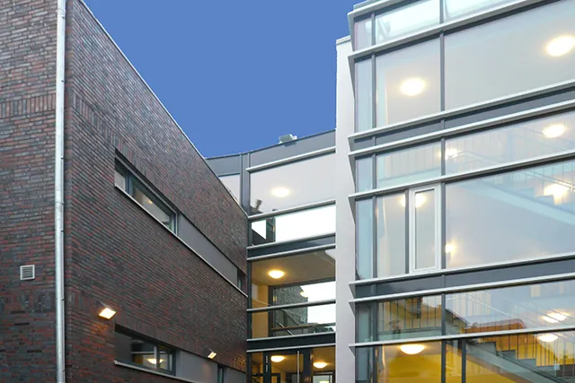 Abendaufnahme eines modernen Gebäudeübergangs mit Glasfassade, der einen beleuchteten Treppenaufgang mit einer dunklen Klinkerwand verbindet.