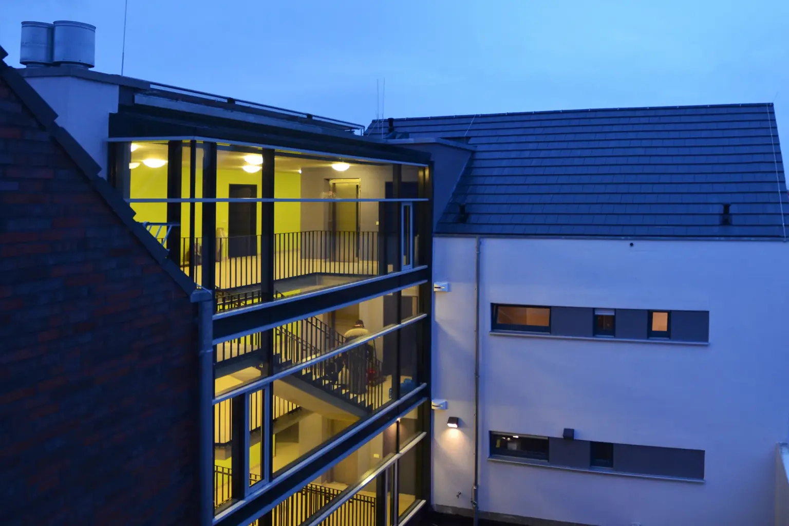 Nachtaufnahme der vollverglasten Treppenhaus-Sektion, die den Blick auf die beleuchteten Flure und die interne Erschließung des Gebäudes freigibt.