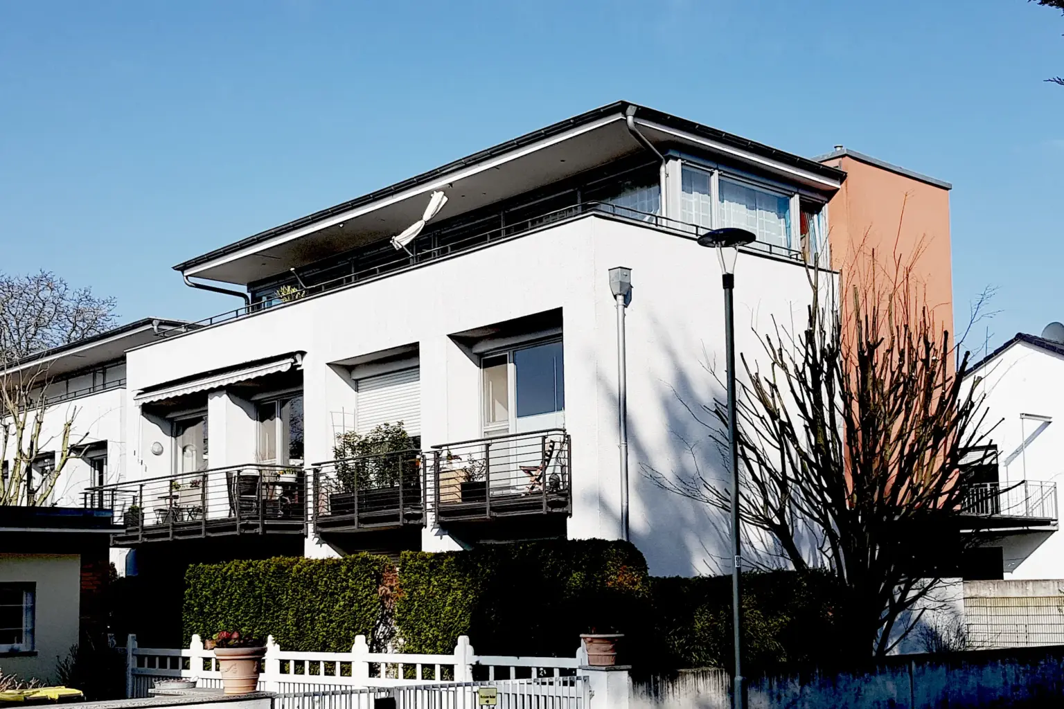 Straßenseite einer Wohnanlage mit großzügigen Balkonen, weißen Fassaden und einem farblich abgesetzten Treppenhaus-Turm in Terracotta.