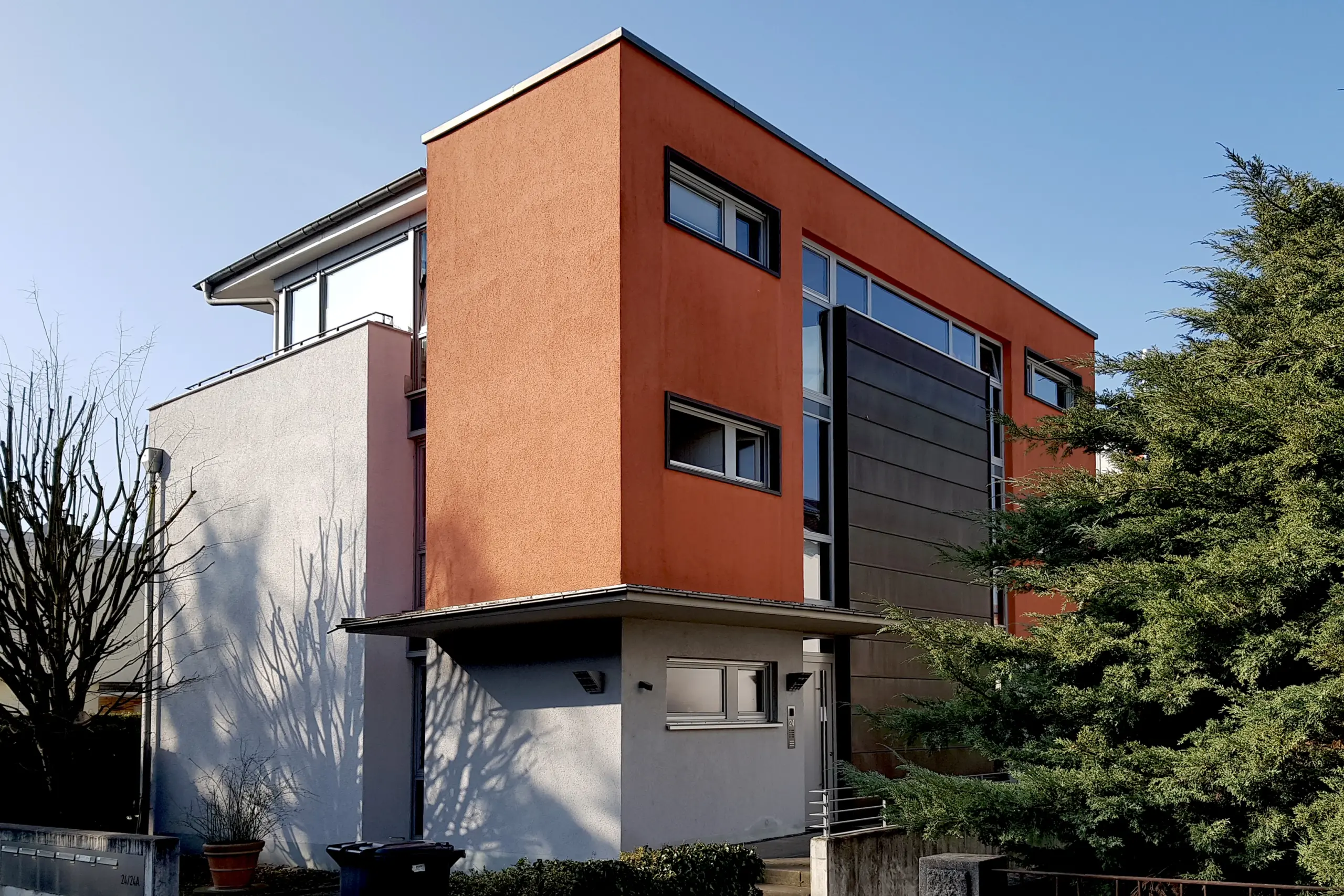 Eckansicht eines modernen Mehrfamilienhauses mit einer Kombination aus terracotta-farbenem und grauem Putz sowie einer dunklen Plattenfassade.