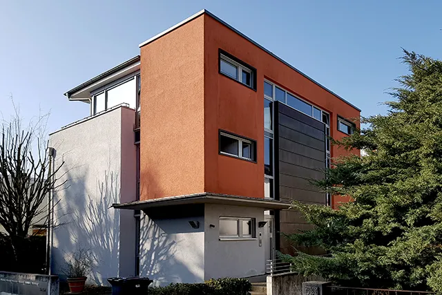 Eckansicht eines modernen Mehrfamilienhauses mit einer Kombination aus terracotta-farbenem und grauem Putz sowie einer dunklen Plattenfassade.