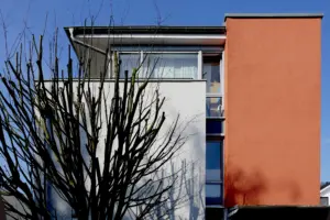 Fassadendetail: Kontrast zwischen weißem Putz und terracotta-farbener Wandfläche, ergänzt durch große Fensterfronten und herbstliche Baumsilhouetten.