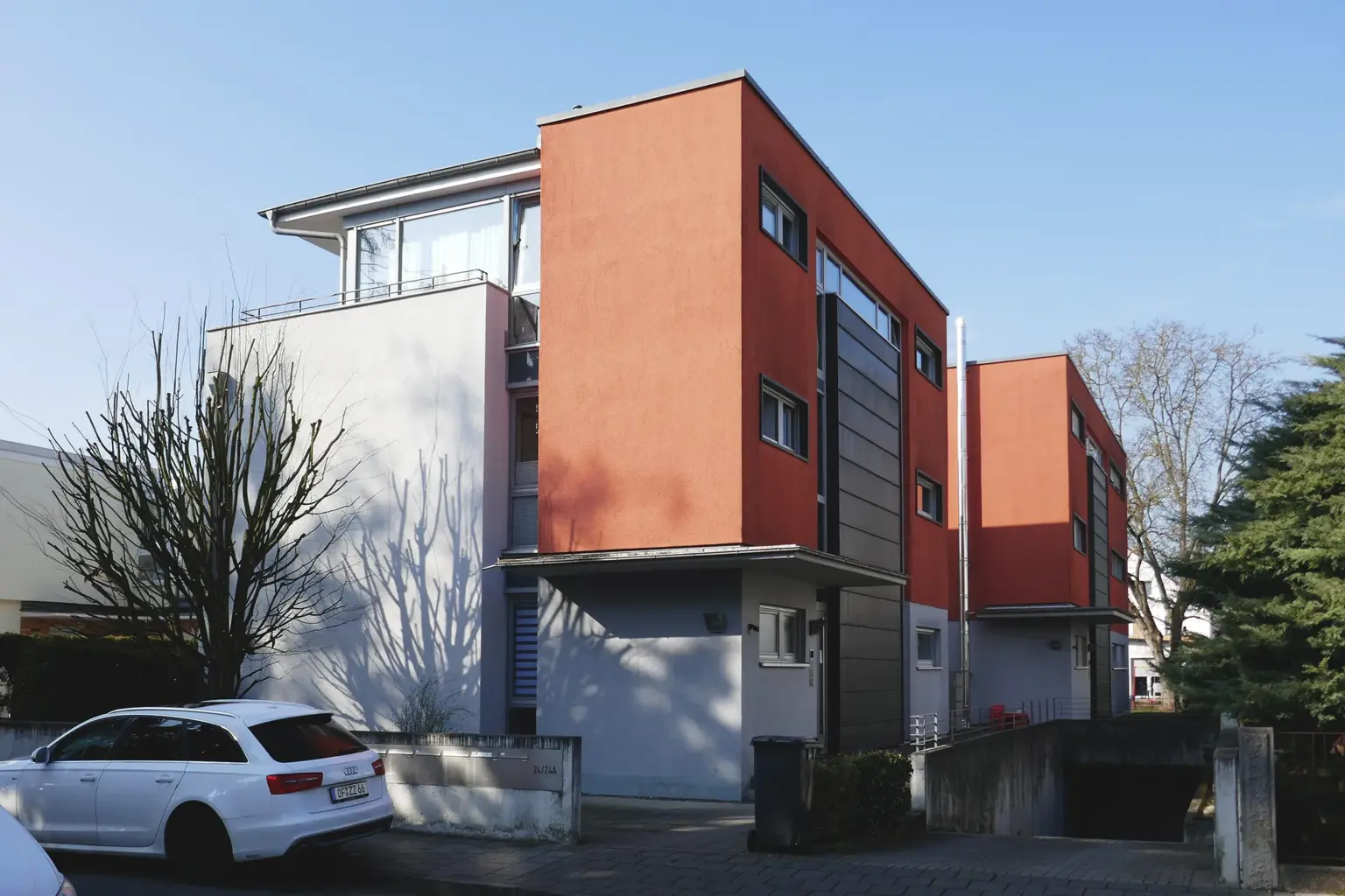 Außenaufnahme zweier moderner Baukörper mit farbiger Fassadengestaltung, Tiefgarageneinfahrt und einem davor geparkten weißen Kombi.