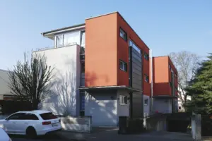 Außenaufnahme zweier moderner Baukörper mit farbiger Fassadengestaltung, Tiefgarageneinfahrt und einem davor geparkten weißen Kombi.