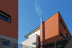 Detailansicht eines glänzenden Edelstahl-Schornsteins an einer modernen terracotta-farbenen Fassade vor strahlend blauem Himmel.