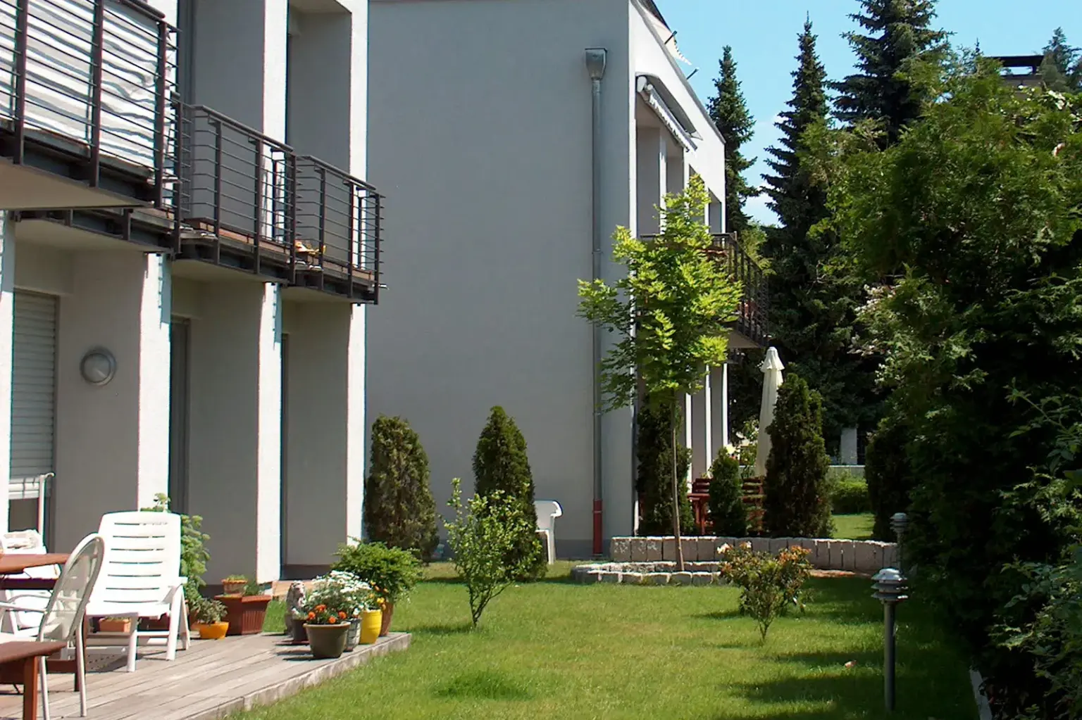 Blick in den Gemeinschaftsgarten einer Wohnanlage mit gepflegtem Rasen, kleinen Nadelbäumen und weißen Terrassenmöbeln.
