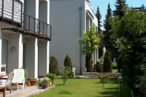 Blick in den Gemeinschaftsgarten einer Wohnanlage mit gepflegtem Rasen, kleinen Nadelbäumen und weißen Terrassenmöbeln.