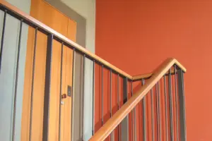 Innenansicht eines Treppenhauses mit einer hölzernen Wohnungstür, Metallgeländer und einer kräftig orange-roten Wandgestaltung.