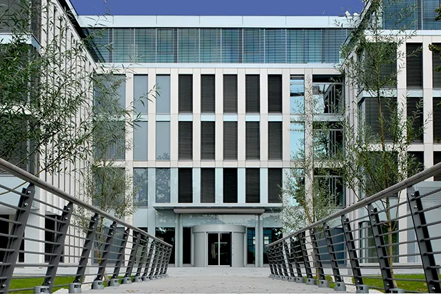 Symmetrische Frontansicht eines modernen Bürogebäudes mit Glasfassade und Außenjalousien, erreichbar über eine stählerne Fußgängerbrücke.