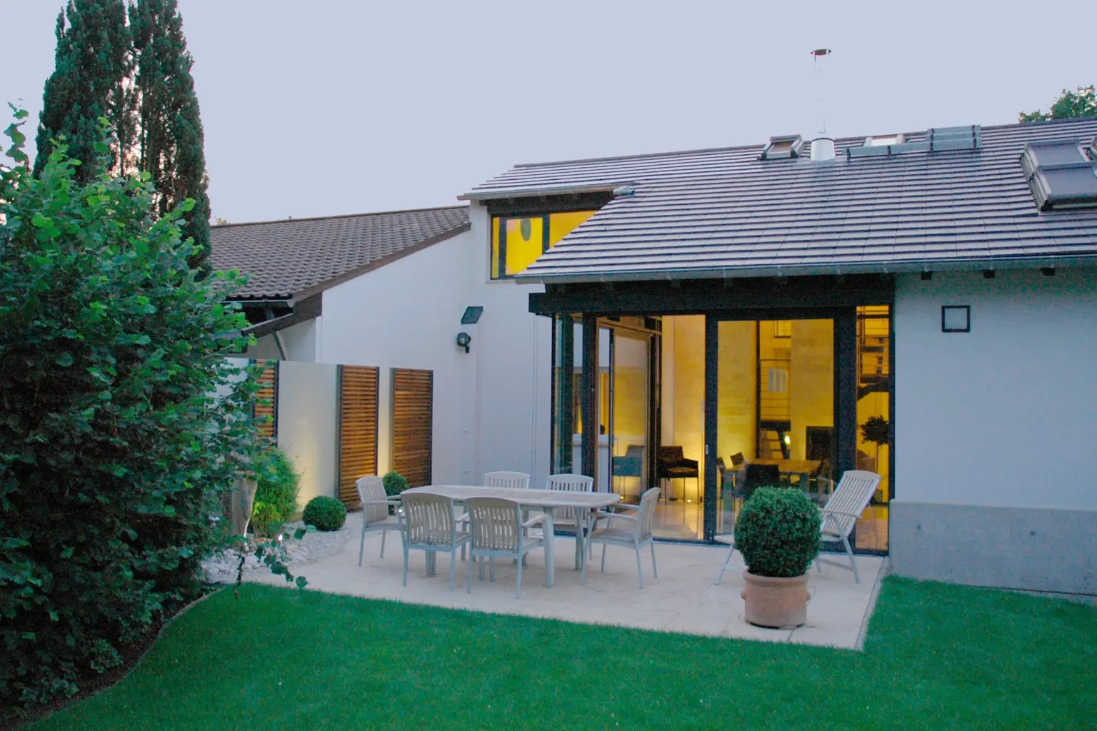 Abendliche Terrassenansicht eines Wohnhauses mit Satteldach, großer Glasfront, gemütlicher Sitzgruppe und beleuchteten Sichtschutzelementen aus Holz.