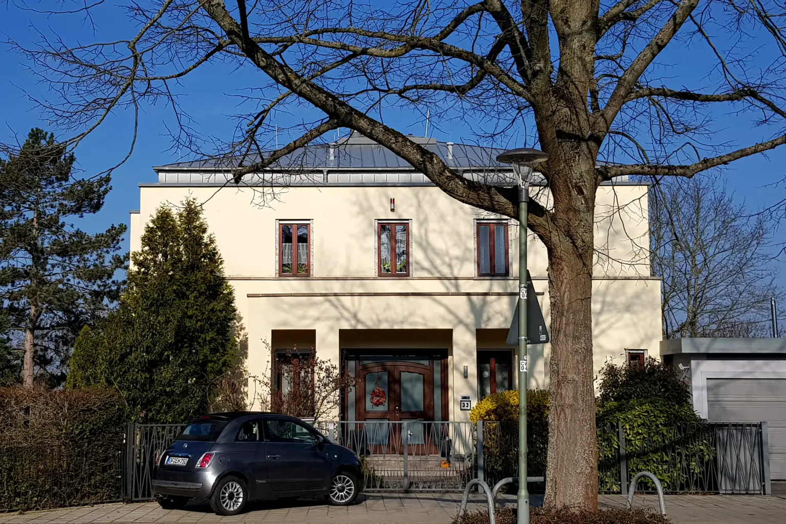 Klassisch-moderne Stadtvilla mit hellgelber Fassade, symmetrischer Fensteranordnung und einem gepflegten Vorgarten mit altem Baumbestand.