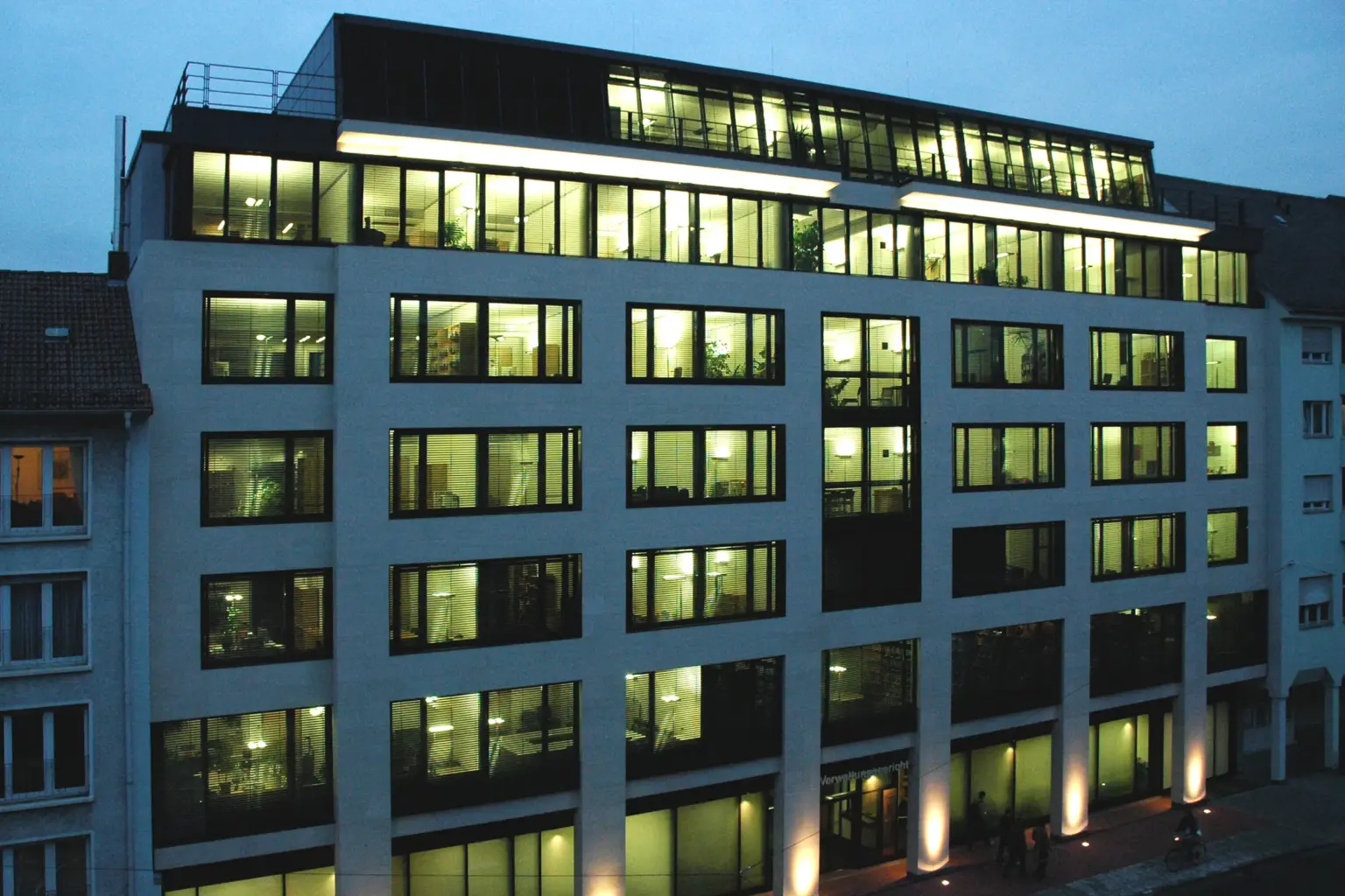 Nachtaufnahme eines mehrstöckigen Verwaltungsgerichts; die beleuchteten Büroräume hinter der Rasterfassade erzeugen ein rhythmisches Lichtspiel.
