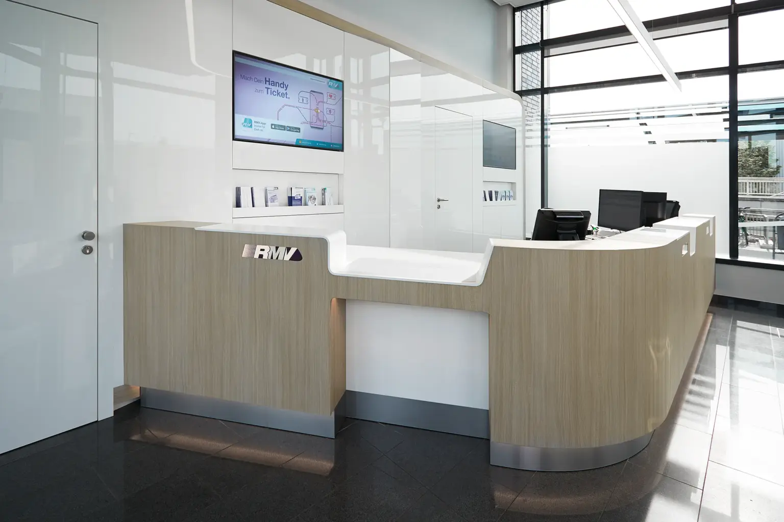 Innenansicht der Mobilitätszentrale: Minimalistischer Service-Tresen in Holzoptik und Weiß vor einer glänzenden Wand mit Monitor-Anzeigen.